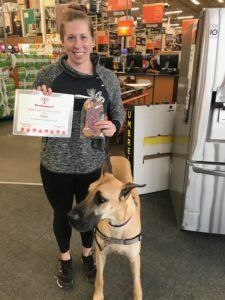 Woman and dog at a store. Woman holds certificate and treat bag, smiling. Dog stands at her feet, wearing a harness.