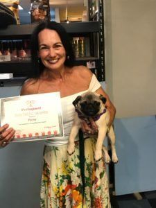 Woman holding a certificate and a pug dog. The woman smiles, wearing a white top and floral skirt.
