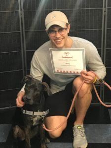 Young man kneels with a dog, holding a certificate. Smiling, wearing a hat, glasses, and a gray shirt.