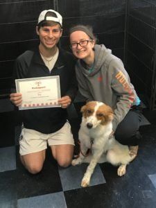 Two people kneel with a dog, holding a certificate. Dark tiled floor, indoor setting.
