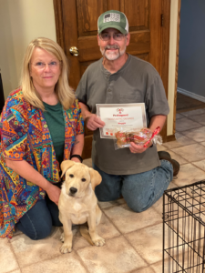 Woman, man, and puppy kneeling, holding certificate and treats, by a crate, indoors.