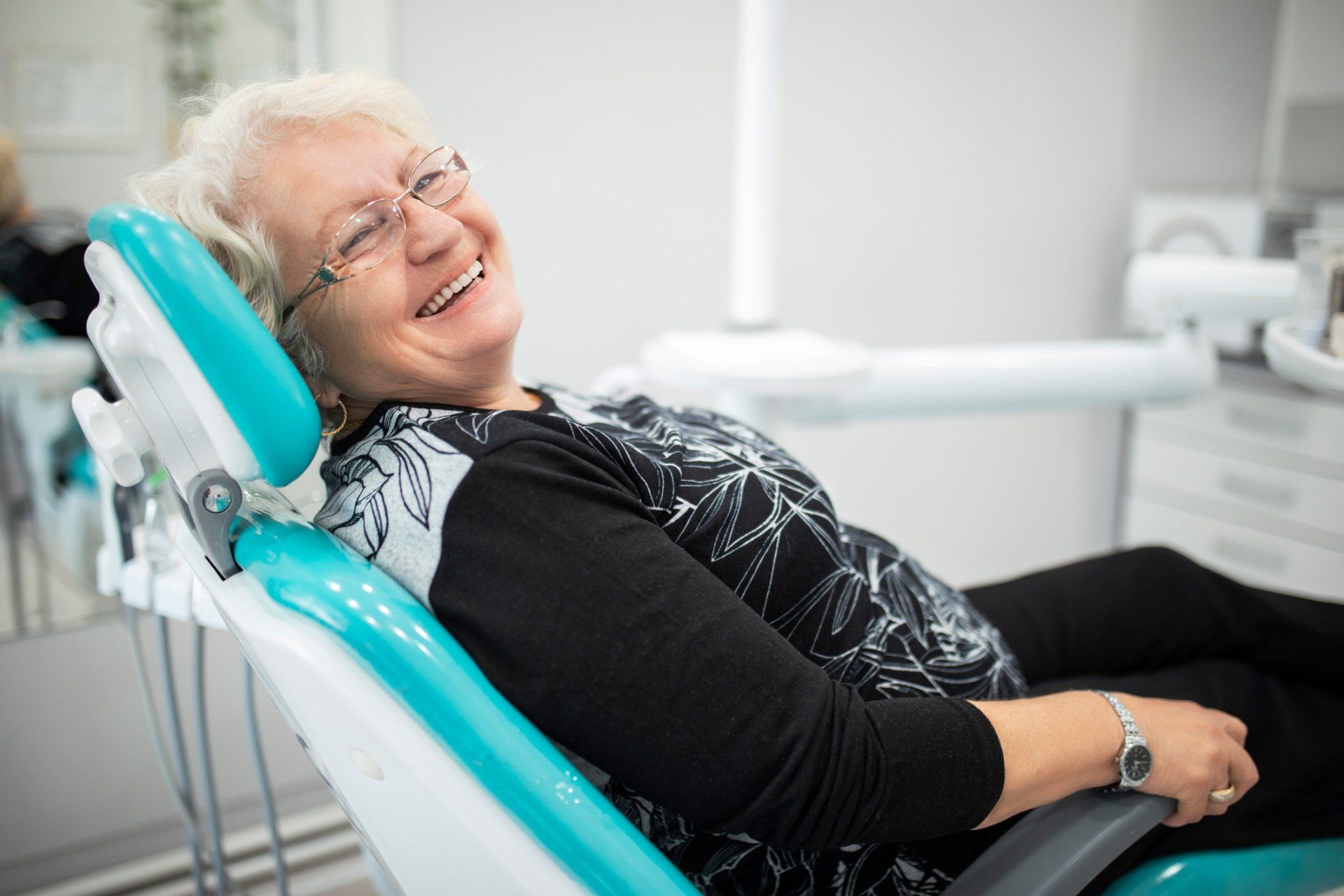 Woman in dental chair, smiling brightly. Turquoise chair, bright setting.