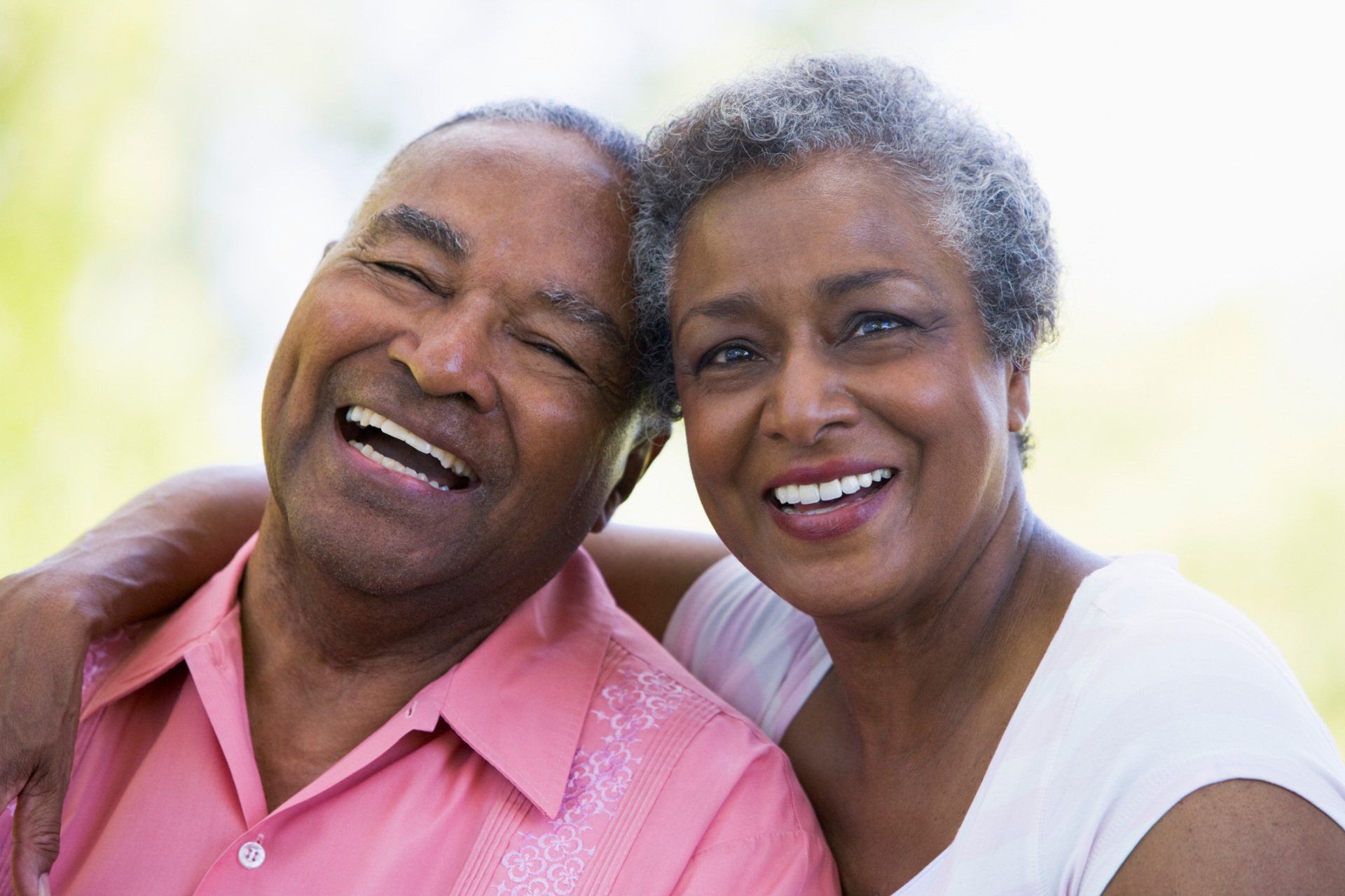 Smiling couple with arm around the other, outdoors; light pink shirt and white top.
