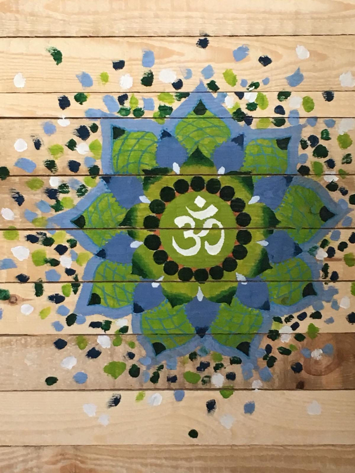 Groene en blauwe mandala met een Om-symbool op een houten achtergrond, omringd door verspreide verfstippen.