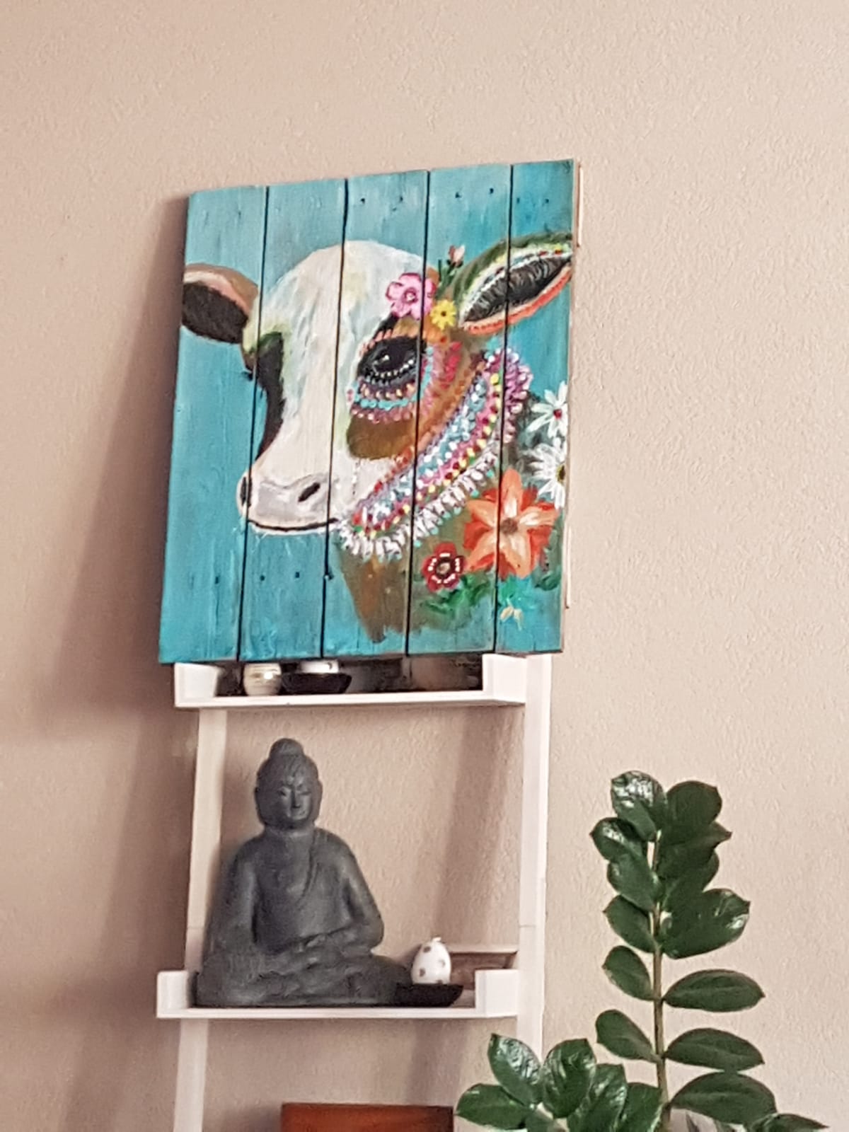 Een turquoise schilderij van een koe hangt boven een witte plank met een Boeddhabeeld en een potplant.