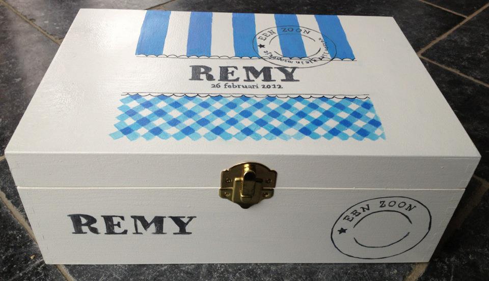 Witte decoratieve doos van het merk REMY met blauwe accenten en een messing sluiting, op een tafelblad.