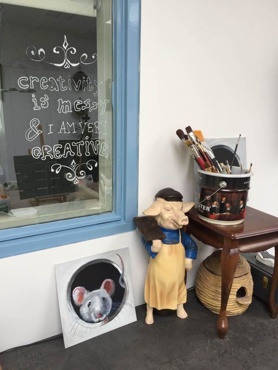 Een kleine etalage met een decoratieve pop, ingelijste kunstwerken en een mand met kwasten