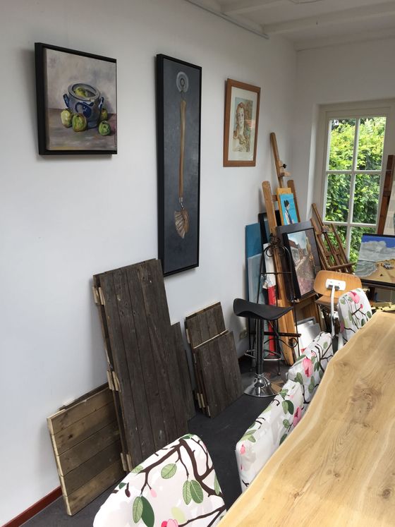 Kunststudio met ingelijste kunstwerken aan de muur, opgestapelde doeken en een lange houten tafel bij een licht raam.