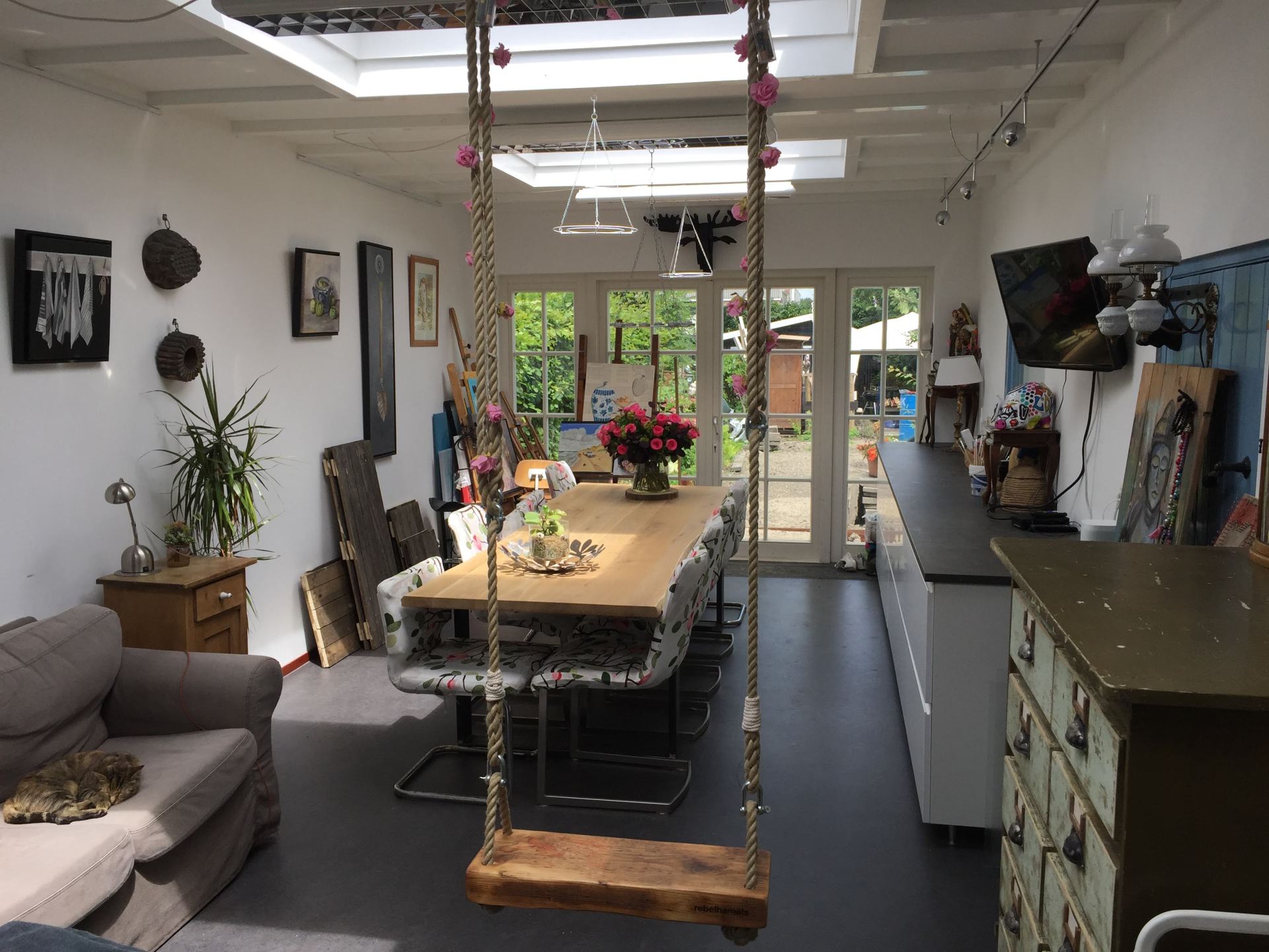 Lichte eetkamer met lange tafel, hangende schommelstoel, planten en openslaande deuren naar de tuin.