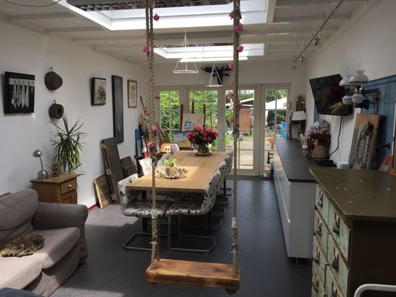 Lichte eetkamer met lange tafel, hangende schommelstoel, planten en openslaande deuren naar de tuin.