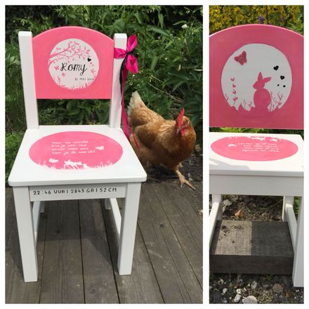 Een witte stoel, roze geverfd met een hartjes- en kattenmotief; ernaast staat een kip op een houten terras.