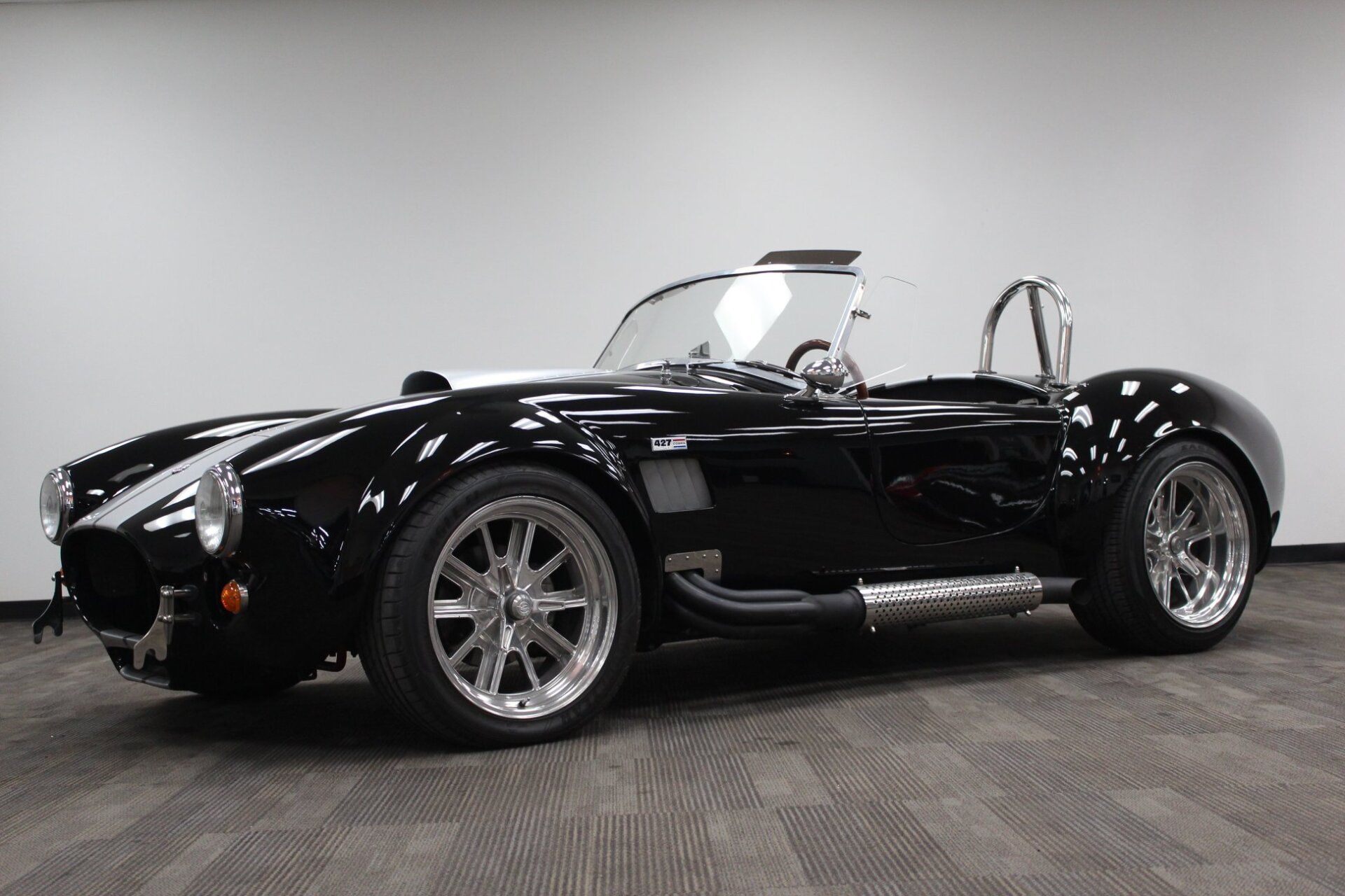 Black classic roadster sports car with chrome accents, parked indoors on a gray floor