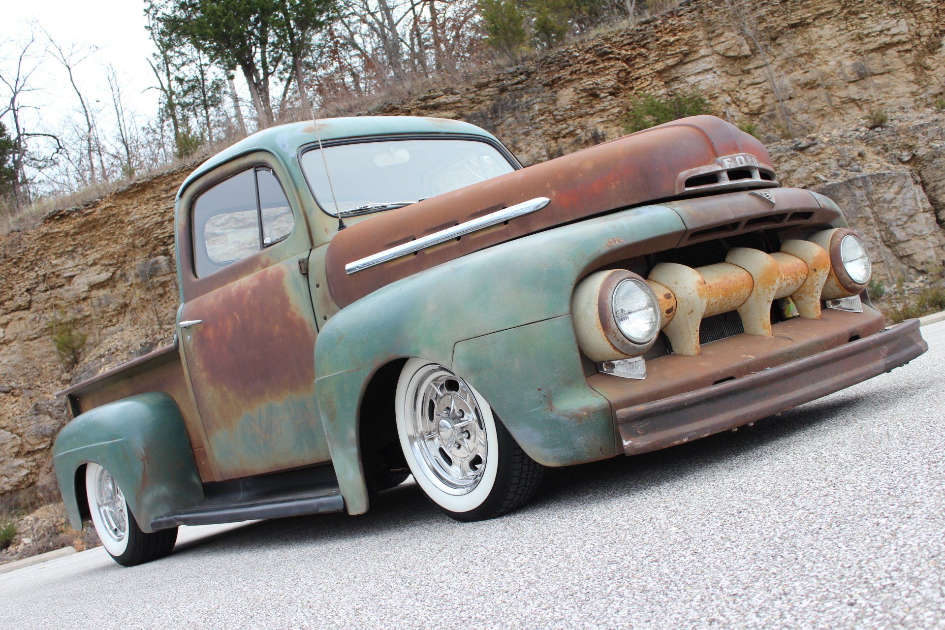 Rusty lowered vintage pickup truck with white rims parked on a roadside hillside.