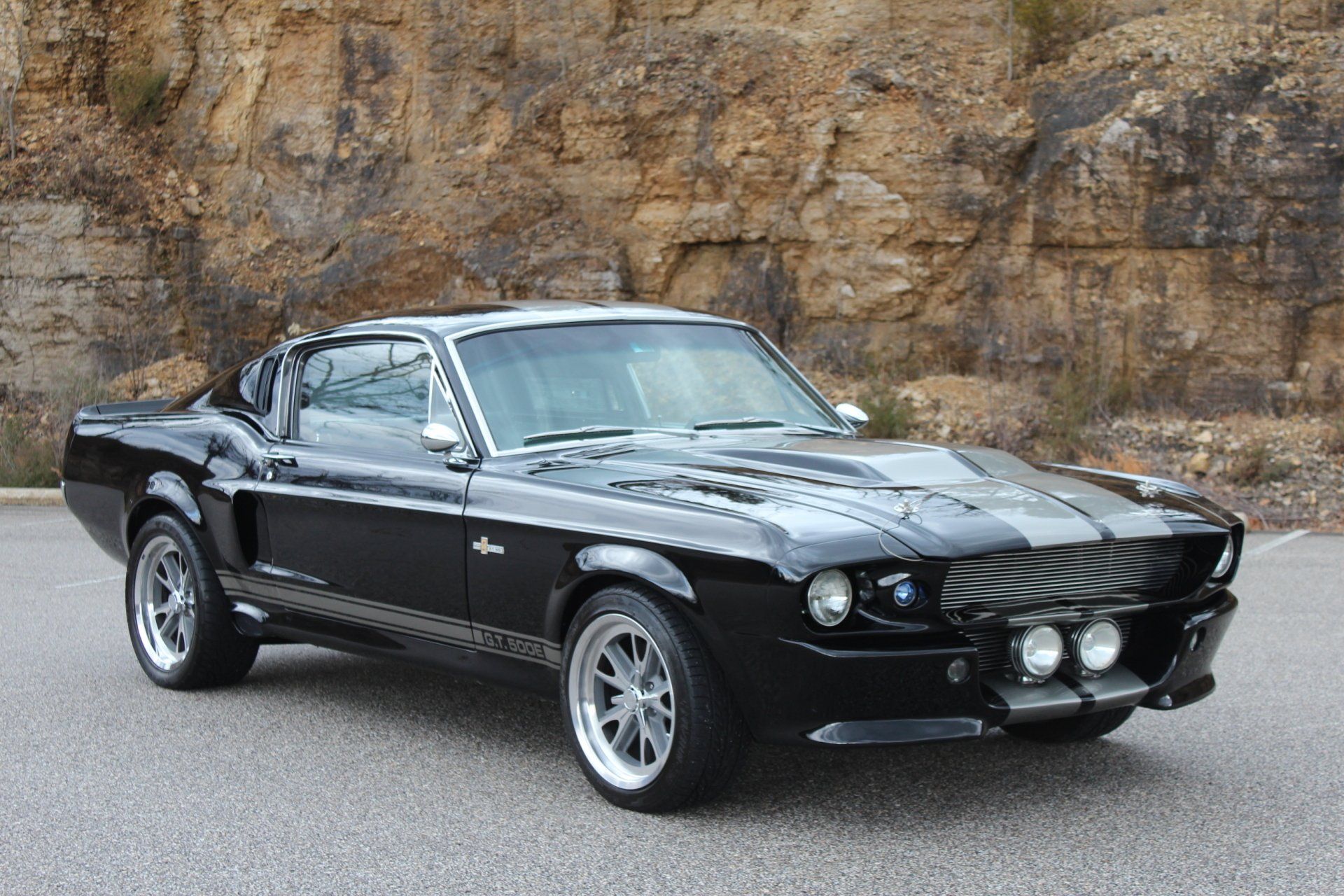 Black classic Ford Mustang coupe parked on pavement in front of a rocky wall
