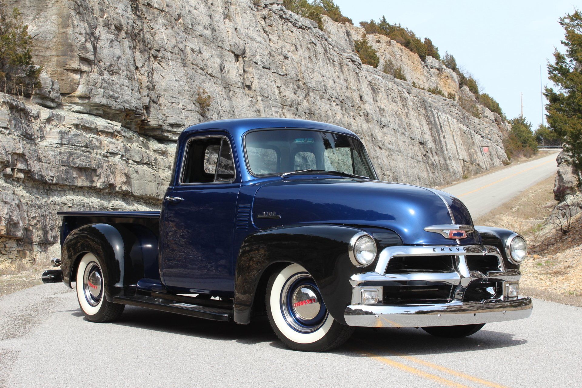 Classic dark blue vintage pickup truck parked on a mountain road beside a rocky cliff.