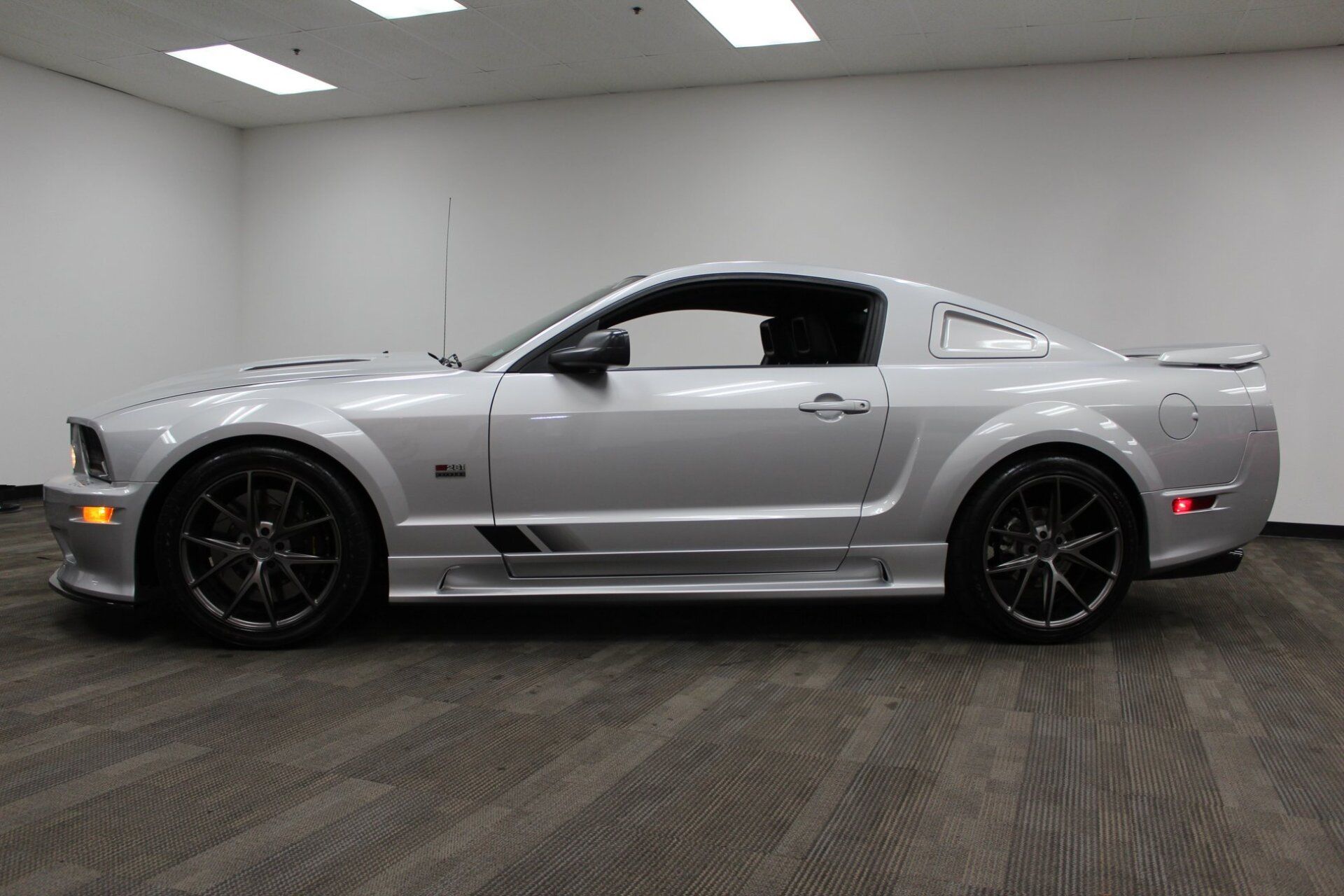 Silver sports coupe in side view, parked indoors on a carpeted floor against a plain gray wall.