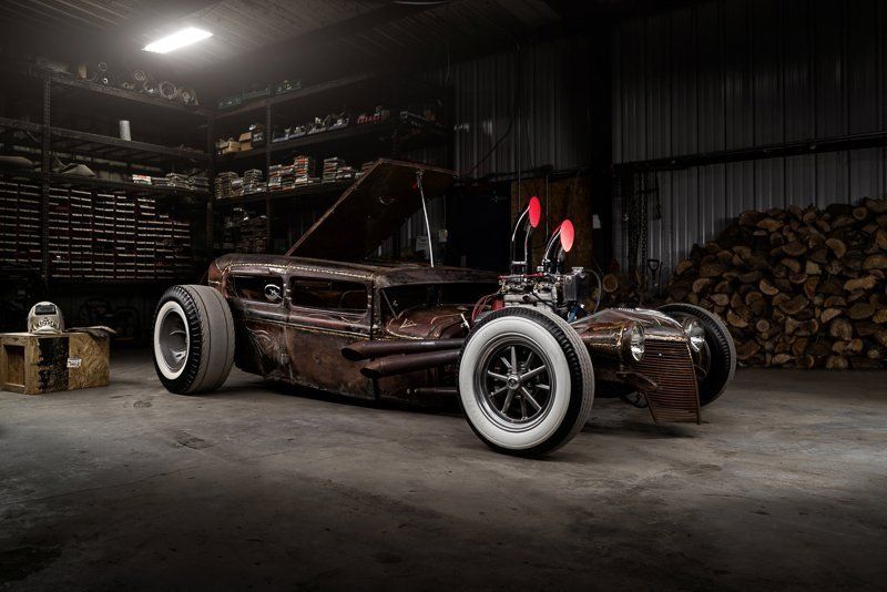 Vintage dark hot rod in a dim garage, with exposed engine and whitewall tires.