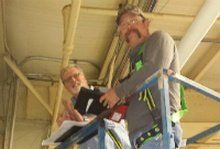Two workers on a lift, inspecting a building ceiling. One holds a clipboard, the other a tool.