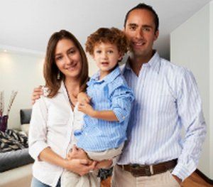 Family of three smiling in a home setting; the mother holds the child.