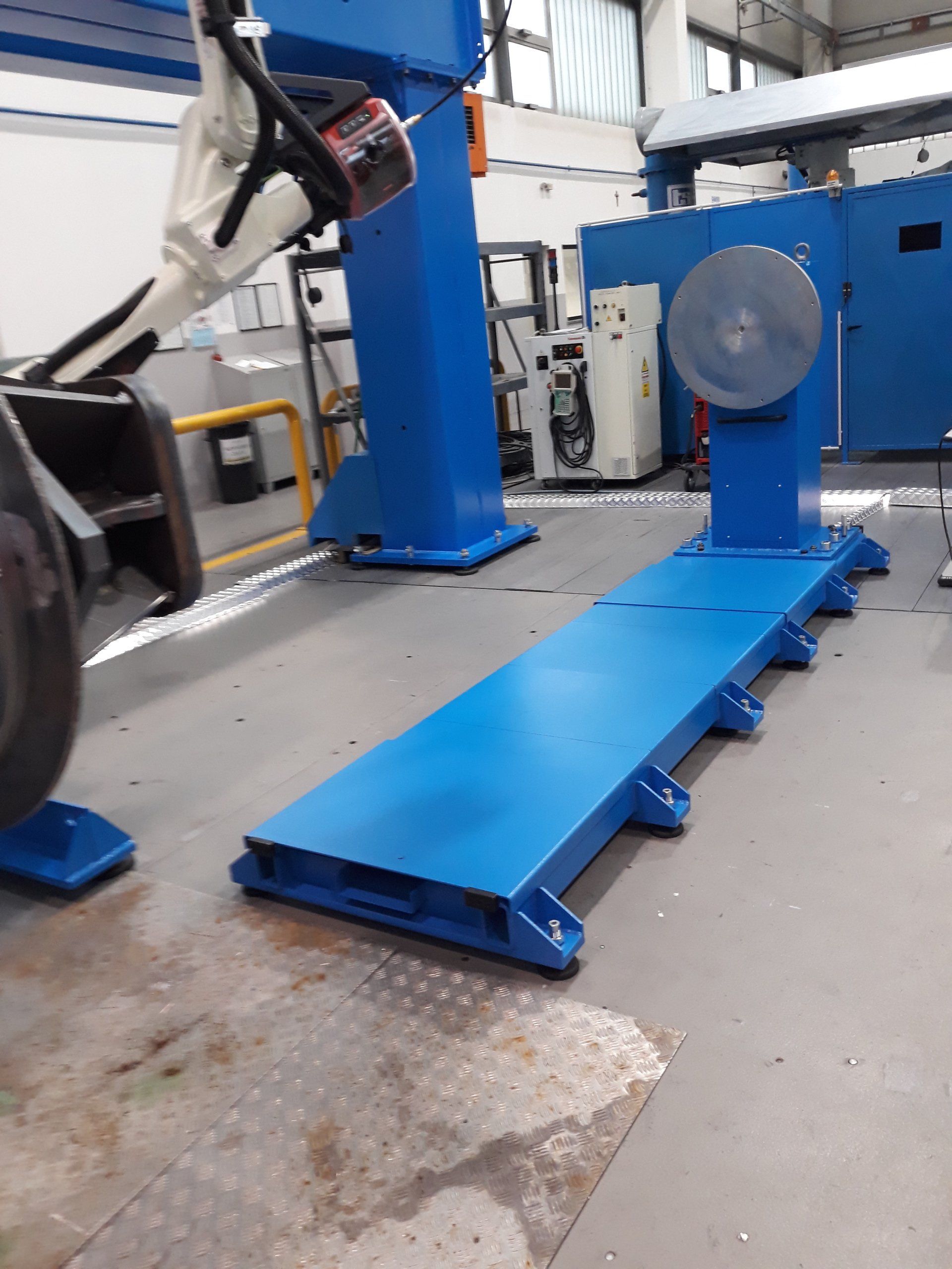 Una cella di saldatura robotizzata industriale con posizionatore di pezzi metallici blu e braccio robotico in un contesto di fabbrica.