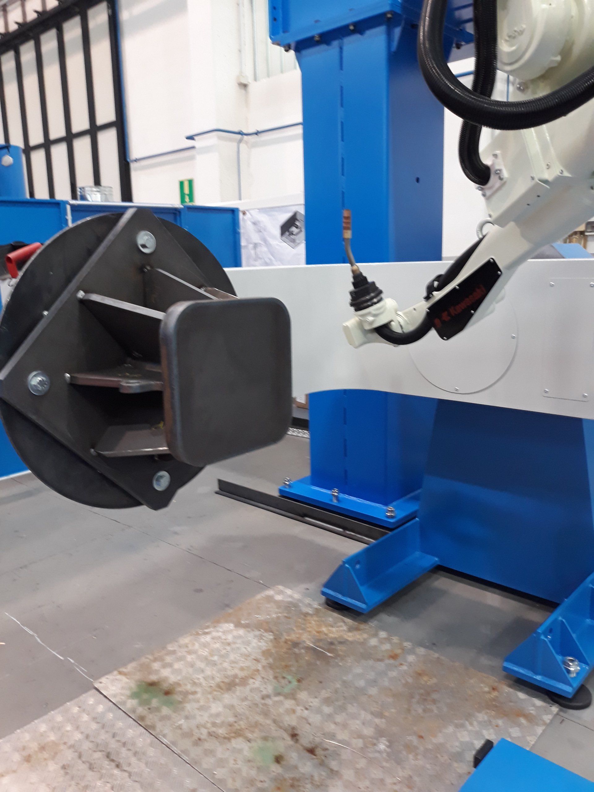 Un braccio robotico posizionato di fronte a un pezzo metallico in lavorazione, fissato su una piastra di montaggio circolare, in un contesto di fabbrica industriale.