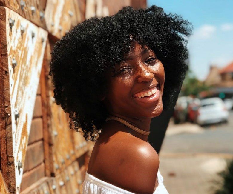 A woman with curly hair is smiling in front of a wooden wall.
