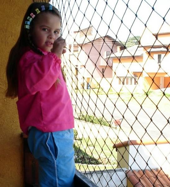 Criança com camisa rosa e calça azul em pé ao lado de uma tela de proteção, olhando para as casas lá fora.