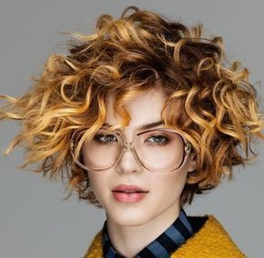 Donna con capelli castani e biondi ricci e scalati, che indossa occhiali e una giacca gialla.