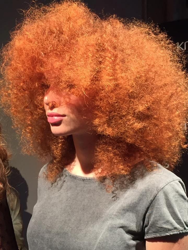 Donna con un grande afro arancione acceso, che indossa un top grigio, illuminata dalla luce del sole.