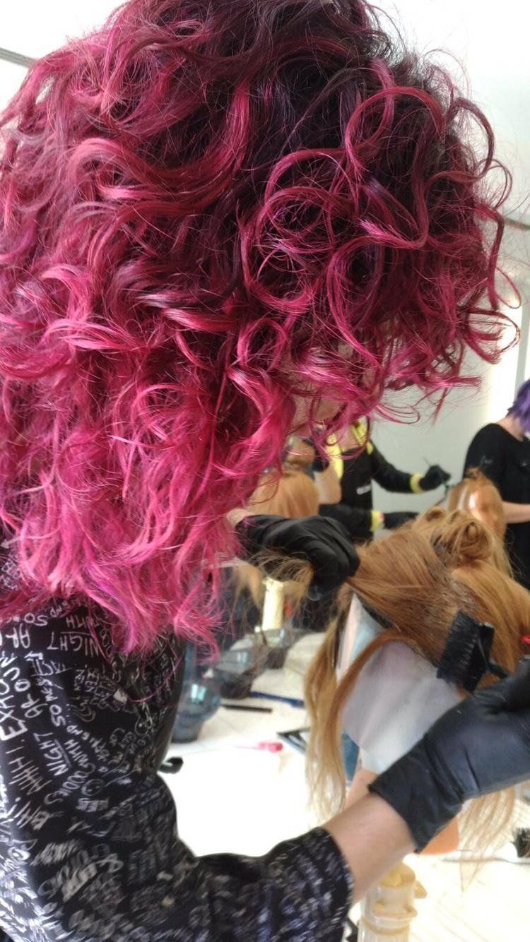 Una persona con i capelli ricci color magenta lavora sui capelli biondi, indossando guanti neri.