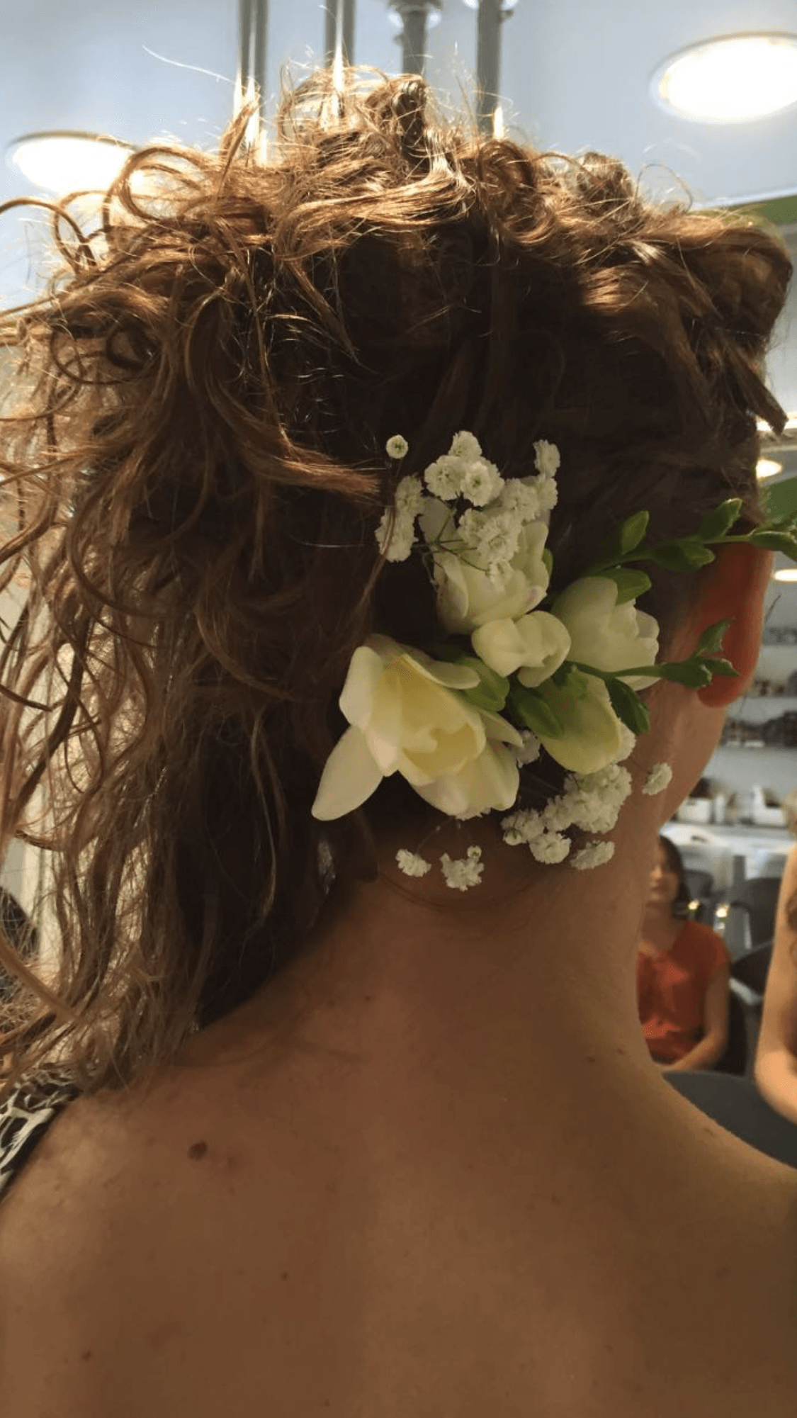 Acconciatura di donna con capelli castani ricci e fiori bianchi. Vista posteriore in una stanza ben illuminata.
