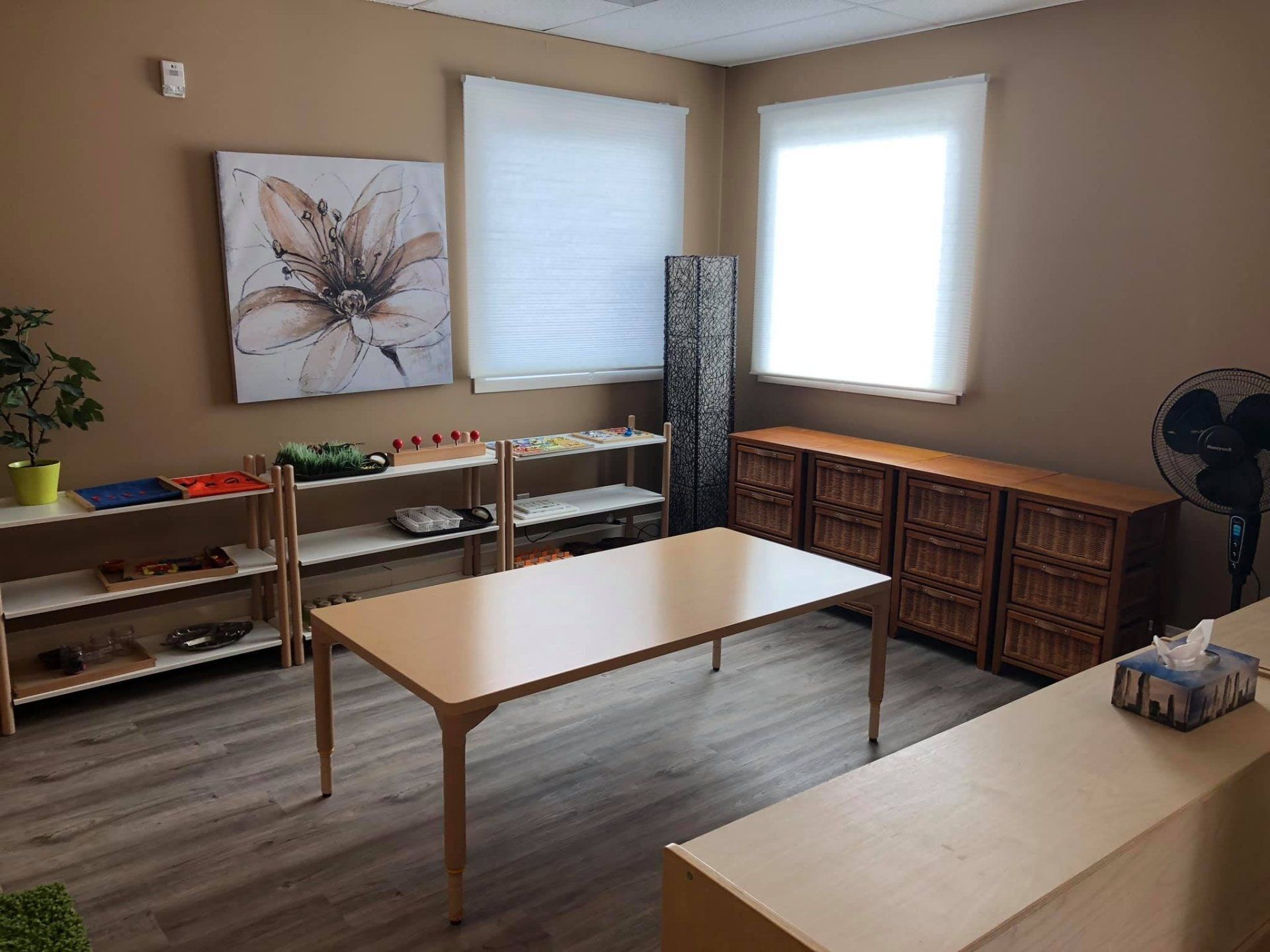 A room with a table , shelves , a fan and a painting on the wall.