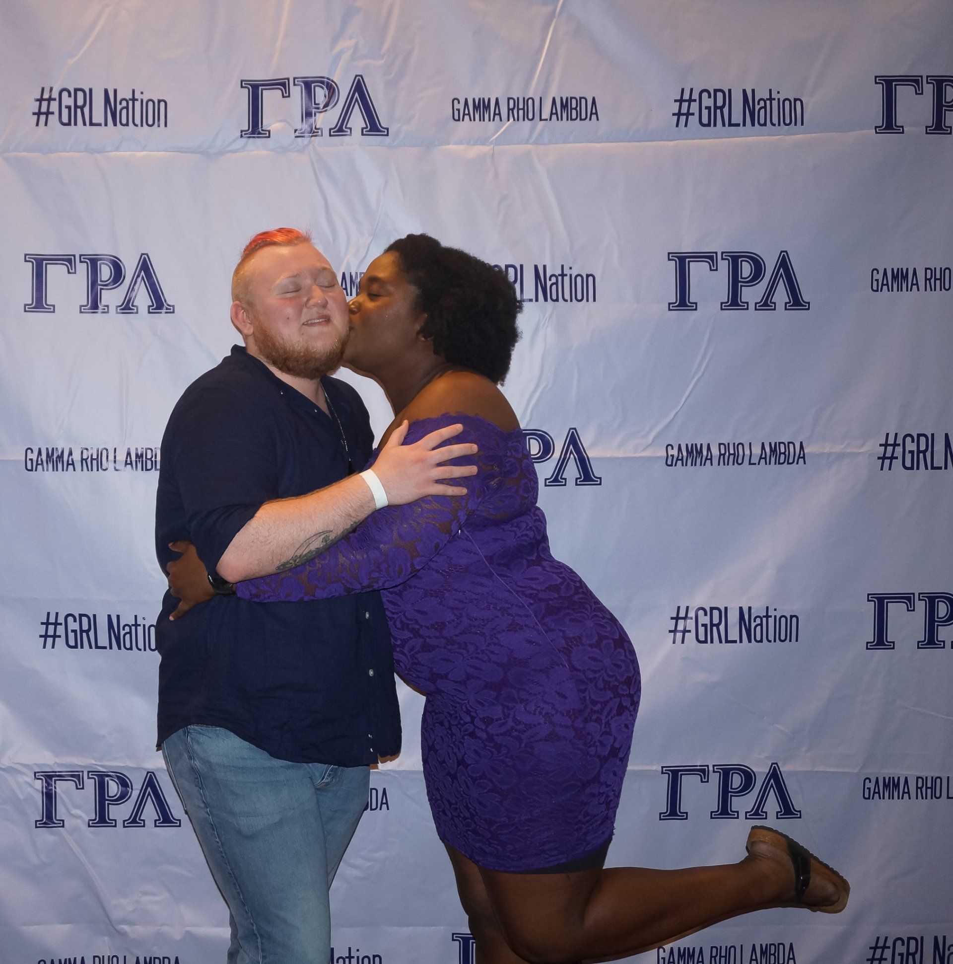 Two people, nicely dressed, stand in front of a background with text on it. The person on the right is kissing the person on the left's cheek