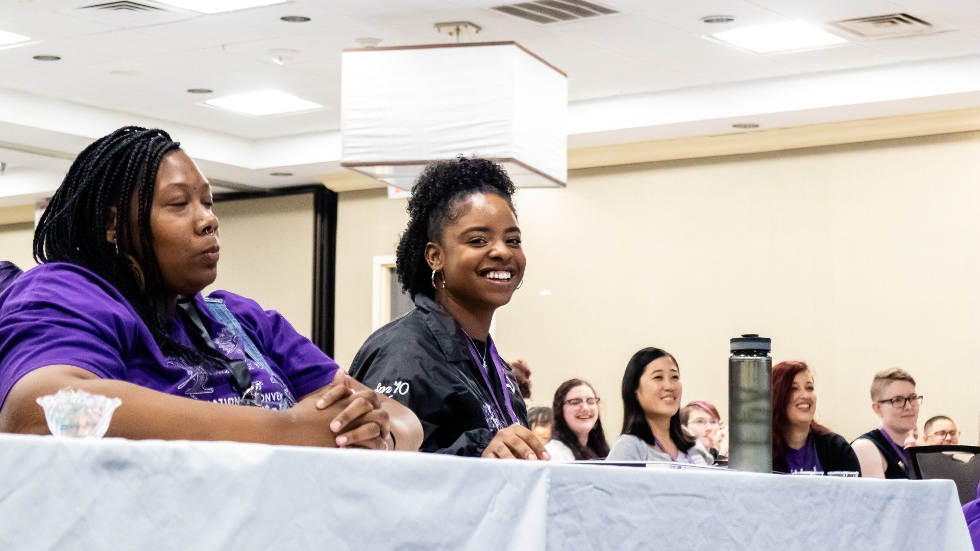 A person smiling at the camera, as others listen to a presentation