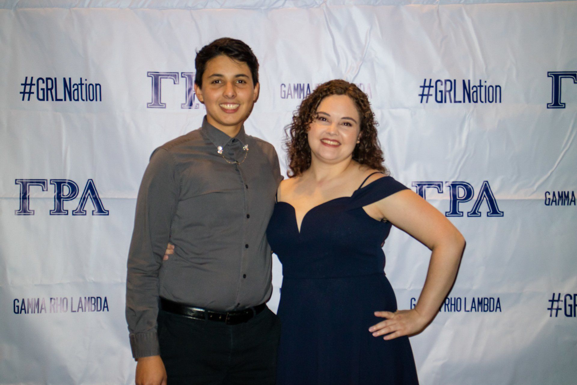 Two people, nicely dressed, stand in front of a background with text on it.