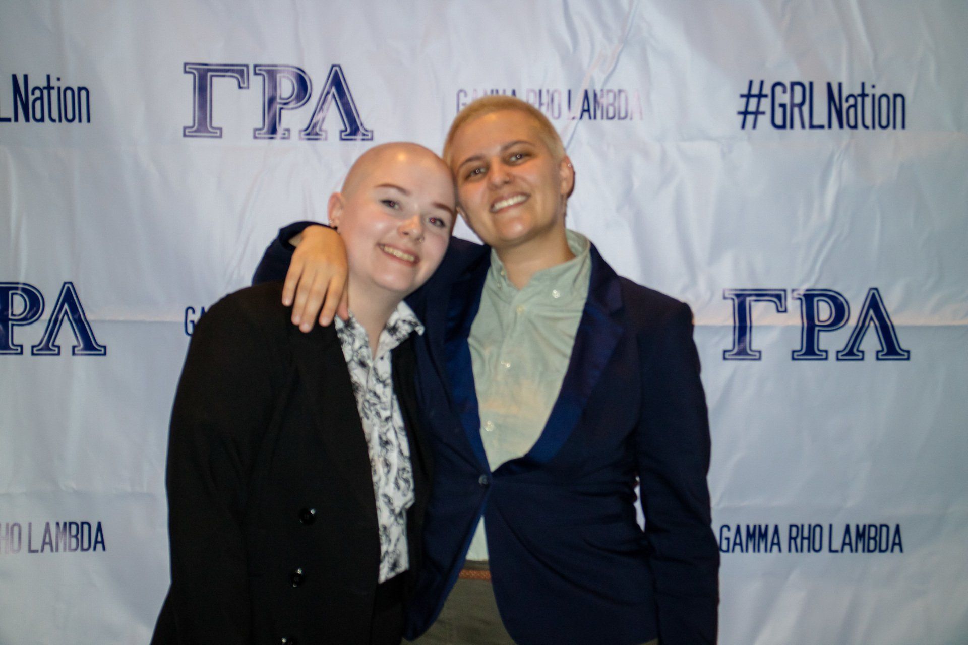 Two people, nicely dressed, stand in front of a background with text on it.