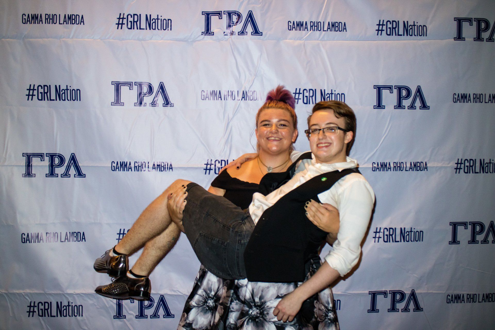 Two people, nicely dressed, pose in front of a background with text on it. One person is carrying the other.