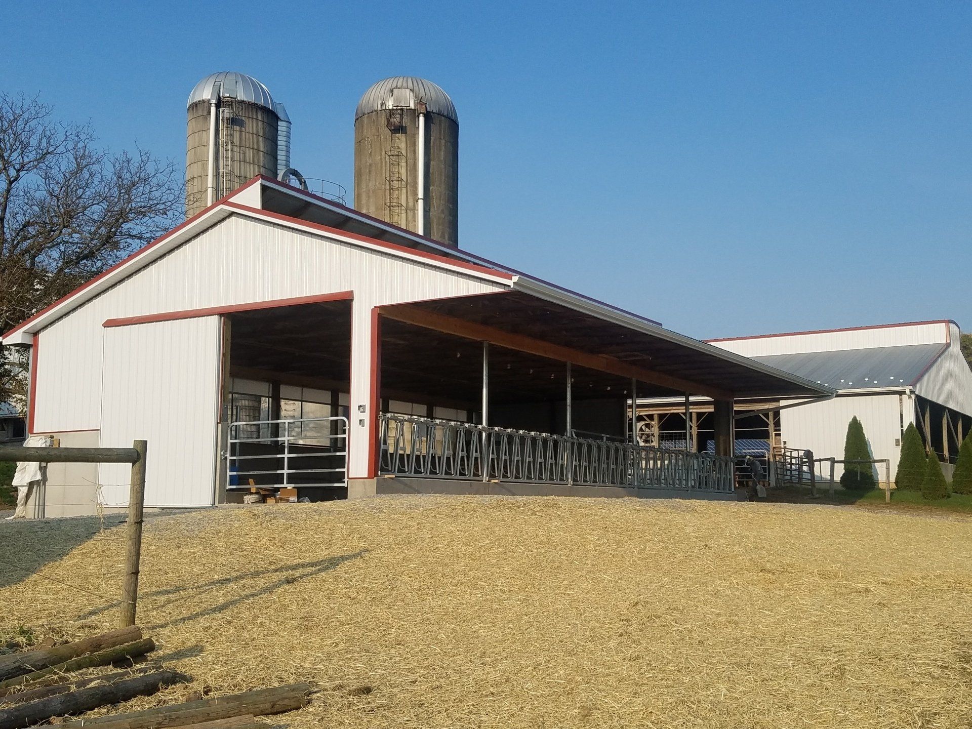 Dairy barn with open-air overhang and two silos under a blue sky, set on a gravel lot.