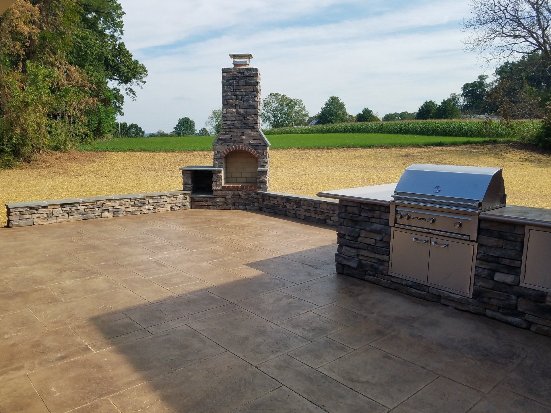Outdoor kitchen with stone fireplace and built-in grill on a concrete patio, fields in the background.