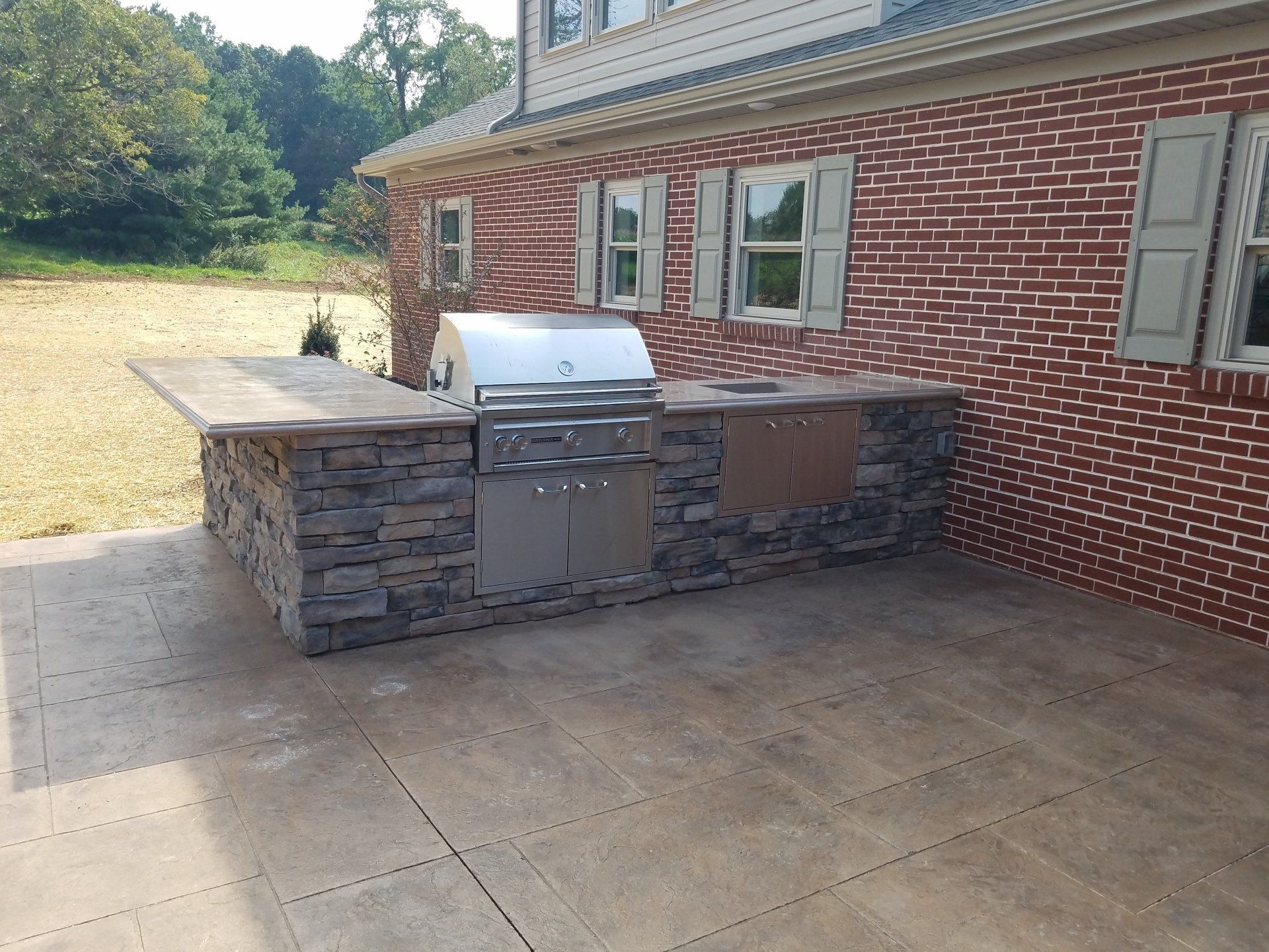 Outdoor kitchen with stone facade, stainless steel grill, and brick house backdrop.