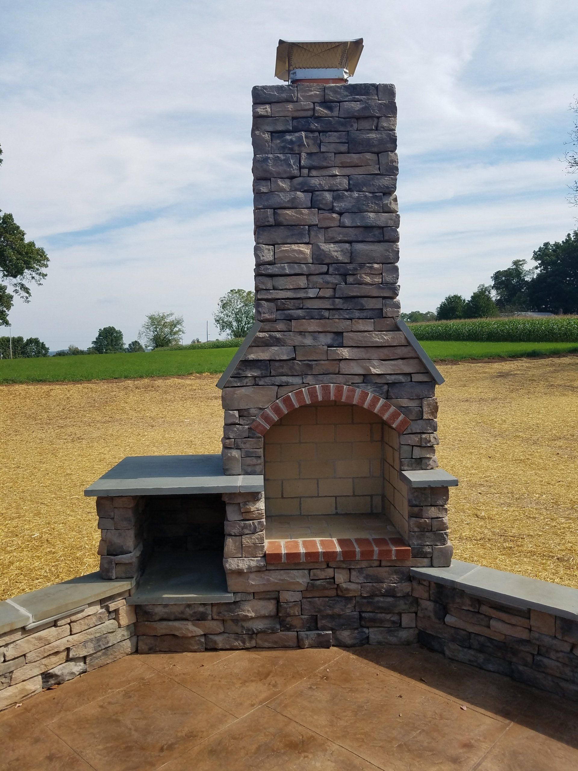 Stone outdoor fireplace with chimney, brick interior, and small side alcove, set in a yard.