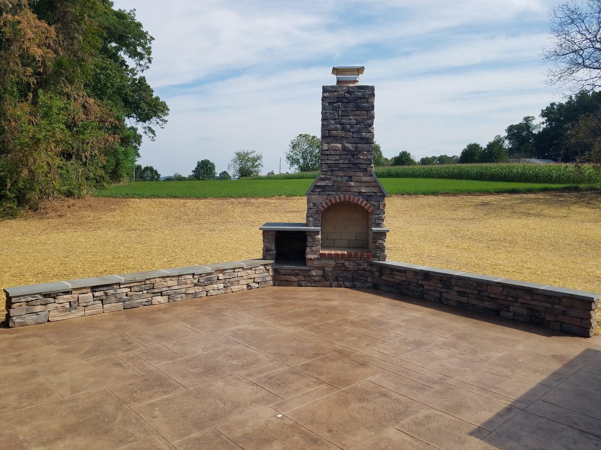 Outdoor stone fireplace with a brick archway, set on a stone patio with surrounding stone walls, fields in the background.