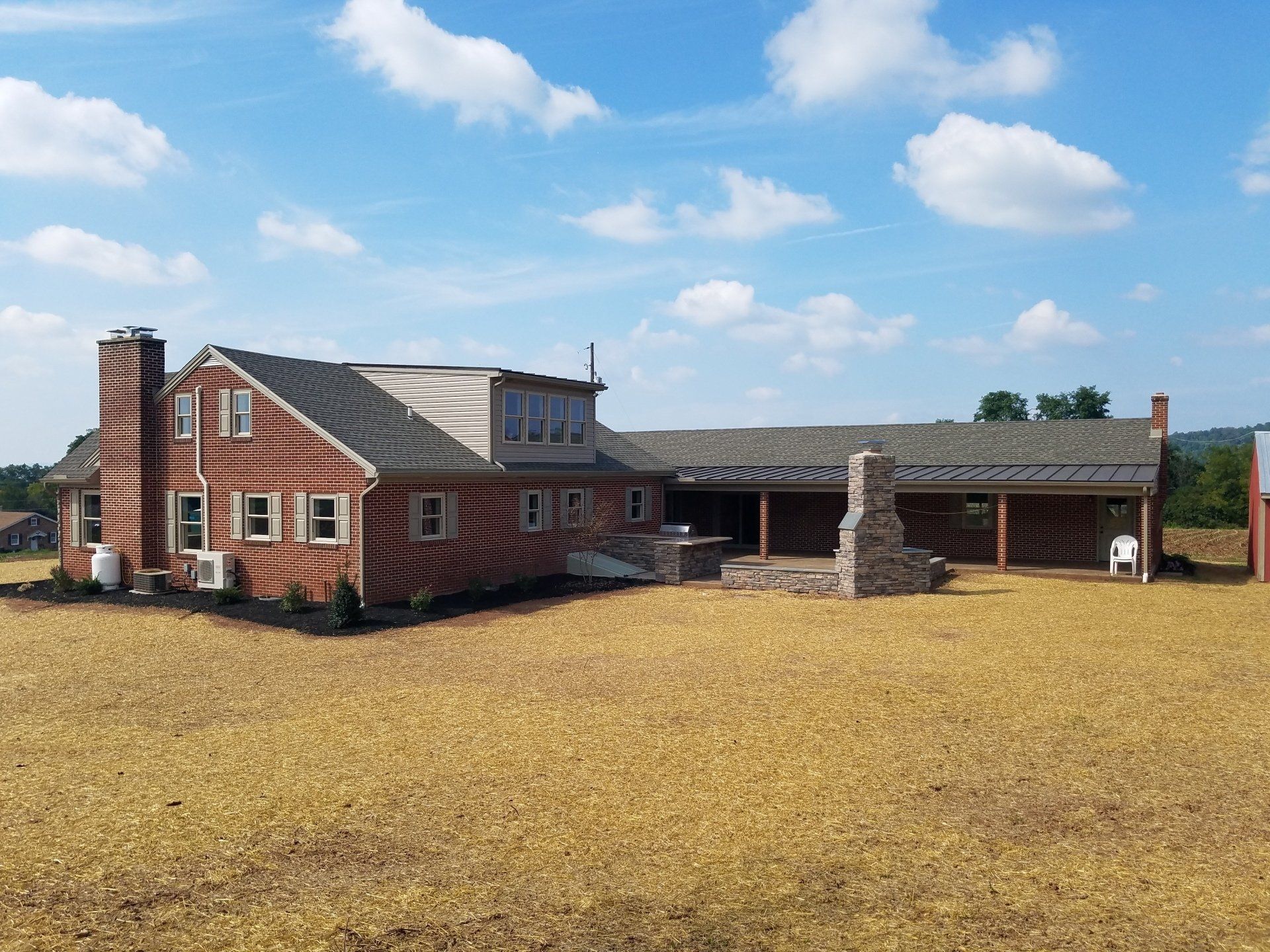Red brick house with a covered patio, set against a field of dry vegetation and a blue sky with clouds.