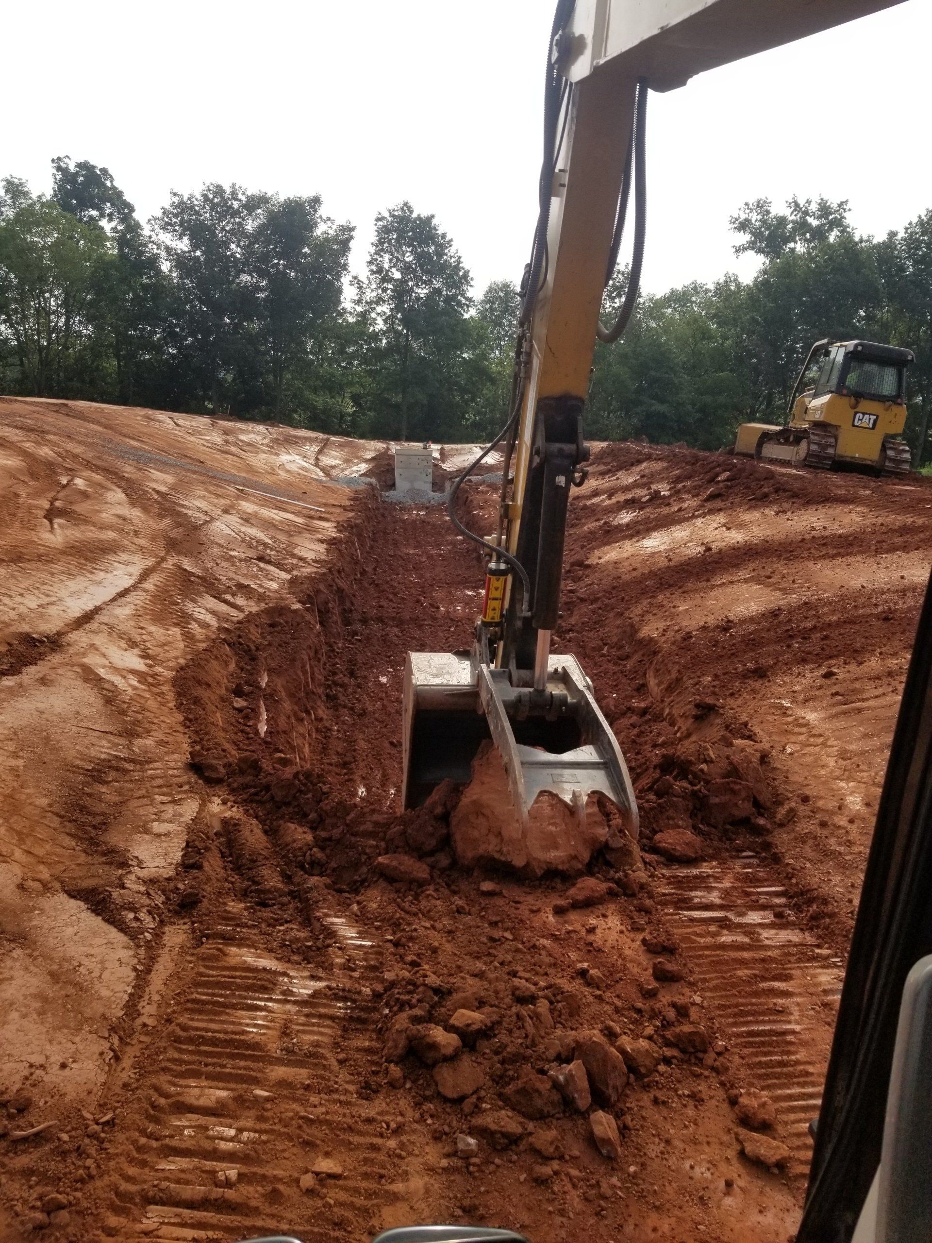 An excavator digging a trench in muddy, red earth. Trees in the background.
