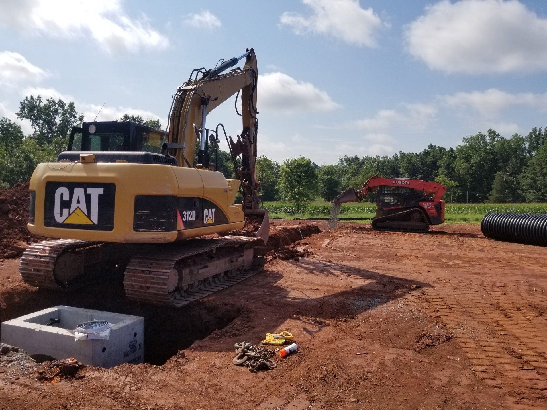 An excavator and a skid steer working on a construction site. Red dirt, concrete box, and black pipes.