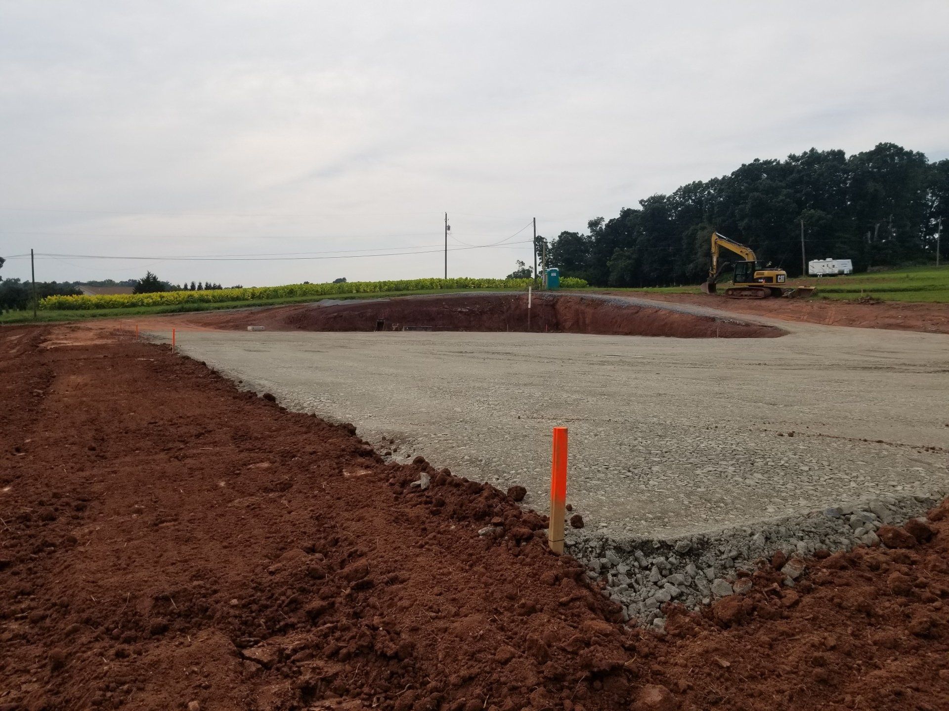 Construction site with gravel foundation and surrounding red soil. Excavator in the distance.