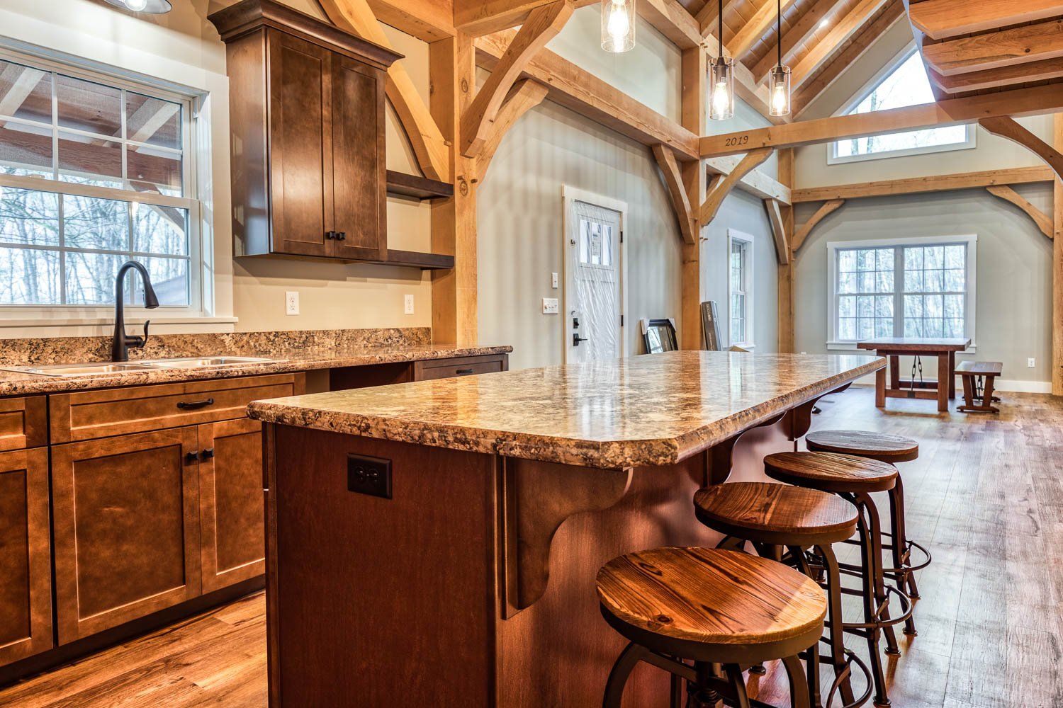 Rustic kitchen with granite countertops, wooden cabinets, and exposed beams; three barstools at island.