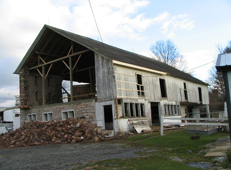 Partially dismantled barn with stone foundation and gray wooden siding against a cloudy sky.