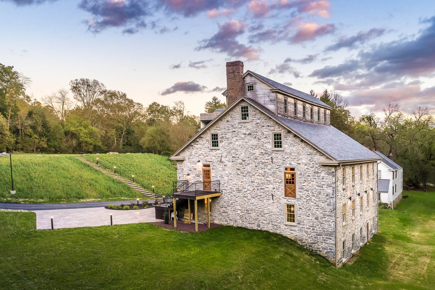 Stone building with a slate roof, a porch, and a grassy yard. Trees and a cloudy sky are in the background.
