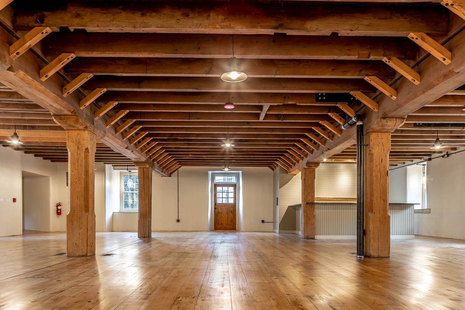 Spacious, empty room with wooden beams and polished wood floor, lit by overhead lights.